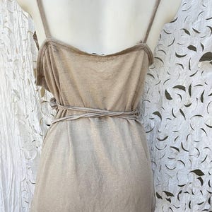 May include: A sleeveless beige tank top featuring thin shoulder straps and a wrap-around tie detail at the waist. The garment is constructed from a solid-colored, lightweight fabric with a scoop neckline and a straight hemline at the bottom.