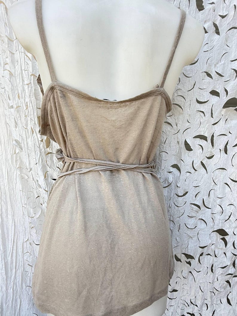 May include: A sleeveless beige tank top featuring thin shoulder straps and a wrap-around tie detail at the waist. The garment is constructed from a solid-colored, lightweight fabric with a scoop neckline and a straight hemline at the bottom.
