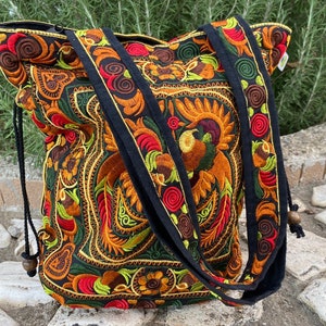 Op de afbeelding: Een zwarte tote bag met een trekkoord. De tas is versierd met kleurrijke geborduurde bloemenpatronen in tinten rood, oranje, geel, groen en bruin.