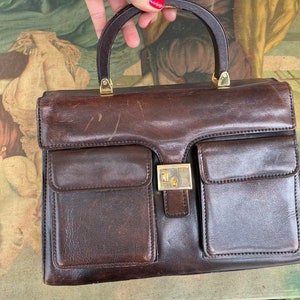May include: A brown leather handbag with a wooden handle and gold accents. The bag has two large pockets on the front and a smaller pocket on the back.