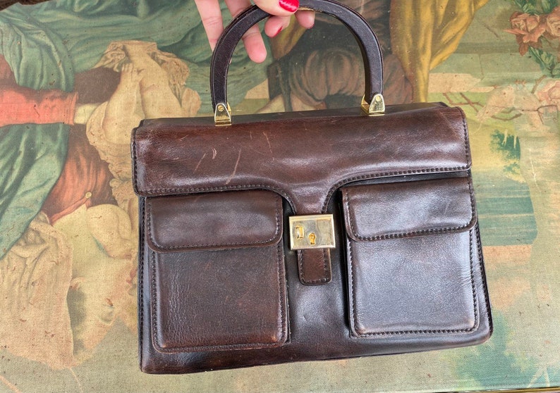May include: A brown leather handbag with a wooden handle and gold accents. The bag has two large pockets on the front and a smaller pocket on the back.