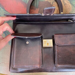 May include: A brown leather handbag with a top handle, a zipper closure, and multiple pockets. The bag has a gold-toned lock and keyhole.