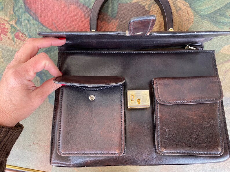 May include: A brown leather handbag with a top handle, a zipper closure, and multiple pockets. The bag has a gold-toned lock and keyhole.
