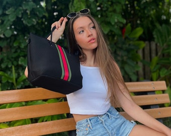 May include: A black tote bag with red and green stripes. The bag is being held by a person sitting on a wooden bench.