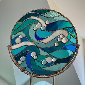 May include: A circular stained glass panel featuring an abstract wave design in various shades of blue, teal, and white. The glass pieces are joined by dark metal solder lines, and the entire piece is framed by a twisted metal border.
