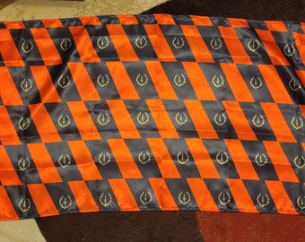Könnte beinhalten: Eine schwarz-orange karierte Flagge mit einem goldenen Lorbeerkranz-Design, das sich über die gesamte Fläche wiederholt.
