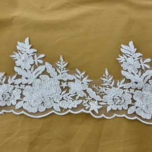 May include: White lace trim with a scalloped edge and floral designs. The lace has a delicate, beaded pattern.