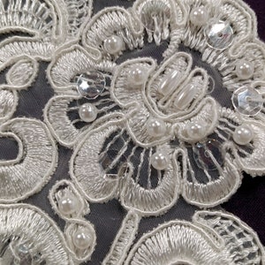 May include: White lace fabric with pearl and sequin embellishments. The lace has a floral design and is delicate and intricate.