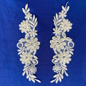 May include: Two white lace appliques with floral designs and pearl accents. The appliques are on a blue background.
