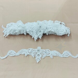 May include: Two pieces of white lace trim with floral and leaf patterns. The top piece is a gathered strip, and the bottom piece is a decorative band with a central embellishment. Both pieces have small, clear embellishments.