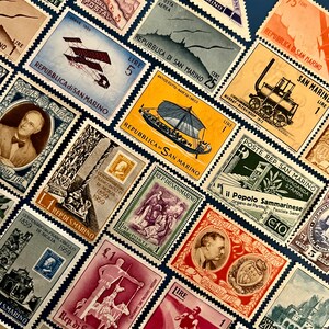 Puede incluir: Una colección de sellos postales antiguos de San Marino, con diversos diseños, incluyendo aviones, figuras históricas y monumentos arquitectónicos. Los sellos son de varios colores y denominaciones, dispuestos sobre una superficie azul oscuro.