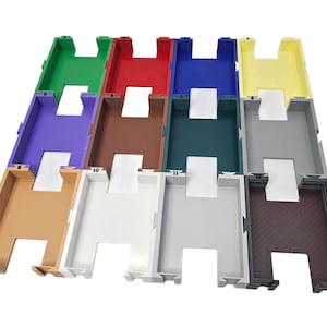 May include: An overhead shot displays a grid of colorful plastic trays. The trays are in various colors, including green, red, blue, yellow, purple, brown, teal, gray, tan, white, light gray, and dark brown. Each tray has a rectangular shape with a central cutout.