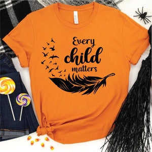 May include: Orange t-shirt with the text "Every child matters" in black script. A black feather and flock of birds design is on the front. The shirt is folded, with a pair of jeans and candy visible in the background.