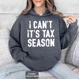 May include: Dark heather gray hoodie with the text "I CAN'T IT'S TAX SEASON" in white block letters. The hoodie has a drawstring hood and a front pocket. The bottom of the image has the text "DARK HEATHER".