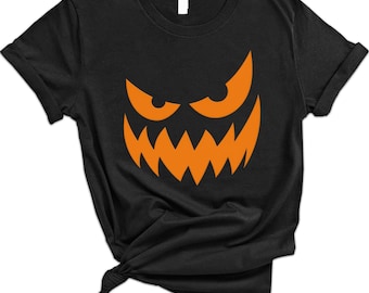 May include: Black t-shirt featuring a jack-o'-lantern face design in orange. The graphic includes angled eyes and a jagged mouth, creating a spooky Halloween theme. The shirt is made of a soft material and has short sleeves.