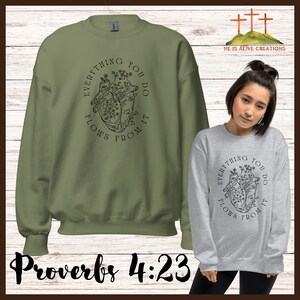 以下が含まれることがあります： 緑と灰色のスウェットシャツに、黒いハートのグラフィックと「あなたがするすべてはそこから流れてくる」と「箴言 4:23」のテキストが書かれています。