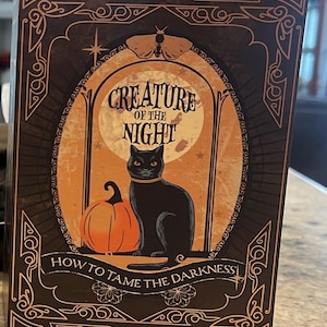 May include: A rectangular book cover featuring a black cat sitting next to an orange pumpkin in front of a full moon. The text reads Creature of the Night and How to Tame the Darkness. The design includes a moth and decorative gold-toned borders.