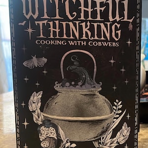 May include: A black book cover titled Witchful Thinking Cooking With Cobwebs in white lettering. The cover features a central illustration of a cauldron with swirling steam, a jar of mushrooms, botanical elements, stars, and a moth.