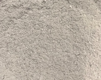 May include: A fine-grained, light gray powder with a dry, matte texture. The substance appears as a dense, uniform layer of loose particles, featuring subtle variations in tone and small, scattered specks throughout the granular surface.