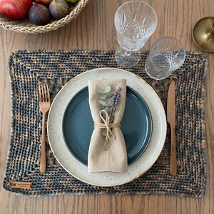 May include: A table setting with a woven placemat, a blue plate, and a beige napkin tied with twine and adorned with lavender. A copper fork and knife are placed on either side of the plate. Two crystal glasses and a lit candle complete the elegant dining scene.