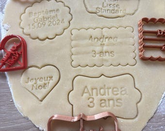 May include: Red and copper cookie cutters on a sheet of dough. The cutters are shaped like hearts, rectangles, and ovals. The dough is cut out with the words "Baptême Gabriel 15.09 2024", "Joyeux Noel", "Andrea 3 ans", and "Andrea 3 ans".