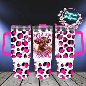 May include: Three stainless steel travel tumblers with pink handles and lids. The tumblers feature a white base with a pink and black leopard print. The center tumbler has a brown cow graphic with the text "JUST A LITTLE MOODY".