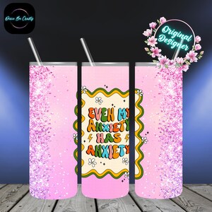 May include: A pink tumbler with a glitter design and the text "EVEN MY ANXIETY HAS ANXIETY" in a retro font. The tumbler includes a silver straw and is decorated with a floral design.