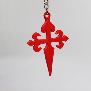 May include: A red, ornate cross with a pointed tip, hanging from a silver chain. The cross is a symbol of the Camino de Santiago, a pilgrimage route to Santiago de Compostela in Spain.