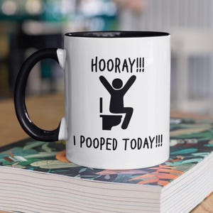 Op de afbeelding: Witte keramische mok met een zwarte handgreep en rand. De mok heeft de tekst "HOORAY!!! I POOPED TODAY!!!" in het zwart, samen met een stokfiguur die op een toilet viert. De mok staat op een boek.