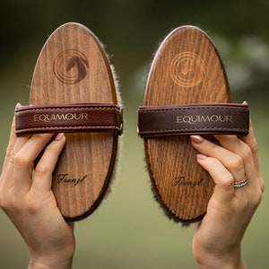 Op de afbeelding: Twee ovale houten poetsborstels met donkerbruine lederen handriemen. Elke borstel is voorzien van de merknaam EQUIMOUR en het woord Franzl in het hout gegraveerd, samen met een cirkelvormig logo van een paardenhoofd. Eén borstel heeft lichte haren.