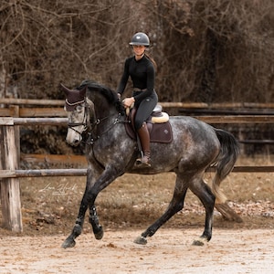 May include: A dapple gray horse with a dark brown bridle, ear bonnet, and saddle pad, ridden by a rider in a black long-sleeve shirt, black breeches, and a black helmet. The horse is moving across a dirt arena with a wooden fence in the background.