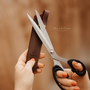 May include: Large, silver scissors with black handles and a brown leather sheath. The scissors are held in front of a blurred background. The sheath has a logo and the word "EASYMOUNT" printed on it.