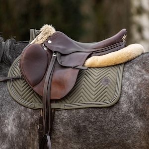 May include: A dark brown leather horse saddle positioned on a grey horse. The saddle features a beige faux fur lining and sits atop an olive green quilted saddle pad with a chevron pattern. Leather stirrup leathers and a metal stirrup are visible.