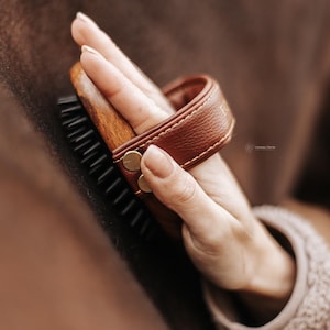 Op de afbeelding: Een houten paardenborstel met zwarte borstelharen en een bruin lederen handvat. De borstel wordt door een hand vastgehouden, wat wijst op verzorging. De afbeelding heeft een warm, natuurlijk kleurenpalet.