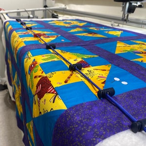 May include: A blue and yellow quilt with a star pattern is being quilted on a longarm quilting machine. The quilt is held in place with blue cords and clips.
