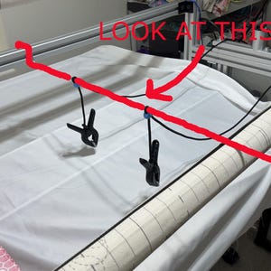 May include: A close-up of a long black rod with two black clips attached to it. The rod is suspended from the ceiling and the clips are holding a white fabric in place. The text "LOOK AT THIS" is written in red on the image.