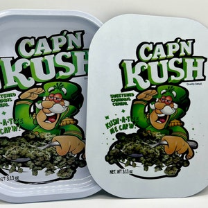 Pode incluir: Uma bandeja de metal branca com um personagem de desenho animado verde e branco. O personagem está usando um chapéu verde e uma jaqueta verde. A bandeja diz "Cap'n Kush" e "Cereal de cannabis adoçado". A bandeja pesa 3,13 onças.