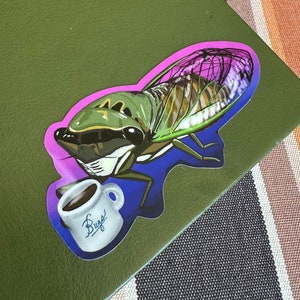 Puede incluir: Una pegatina de una cigarra verde y marrón con detalles en rosa y morado, sosteniendo una taza de café blanca con la palabra "Bug" escrita en ella.