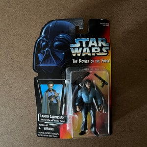 May include: Star Wars The Power of the Force action figure of Lando Calrissian. The figure is wearing a blue shirt and trousers and is holding a blaster pistol. The packaging is blue and red with a picture of Darth Vader on the front.