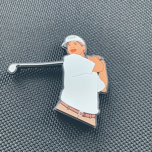 Może przedstawiać: Biała koszulka golfowa z brązowym paskiem i białym kapeluszem z napisem "LYNX". Golfista trzyma kij golfowy i jest w pozycji do uderzenia.