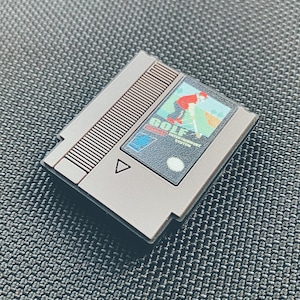 May include: A gray Nintendo Entertainment System game cartridge with a black label that says "GOLF" and features a cartoon golfer swinging a club. The cartridge has a gray plastic shell with a black label and a gray plastic tab.