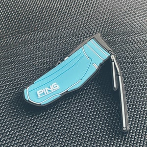 Könnte beinhalten: Ein blauer und schwarzer Golfbag-förmiger Golfball-Marker mit dem Wort "PING" in weißen Buchstaben.