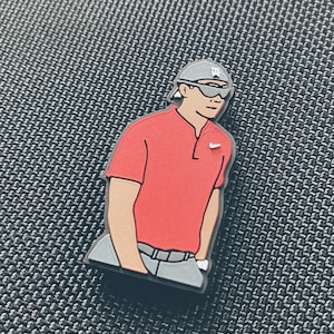 May include: A red golf shirt with a white logo, grey trousers, and a grey baseball cap with a white logo. The figure is wearing sunglasses.