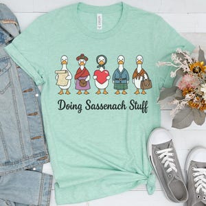 May include: A light green t-shirt featuring a graphic of five cartoon ducks dressed in historical Scottish attire, with the text "Doing Sassenach Stuff" printed in black below the ducks. The shirt has a crew neckline and short sleeves.