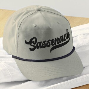 May include: A pale grey baseball cap featuring the word "Sassenach" in black script. The cap has a navy blue rope detail around the brim and a classic, structured design.