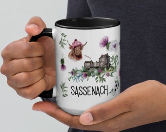 May include: White ceramic mug with a black rim and handle. The mug features a watercolor illustration of a Scottish Highland cow, a castle, and flowers. The text "Sassenach" is printed in black at the bottom of the mug.