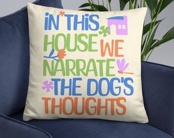 May include: Cream-colored throw pillow with the text "IN THIS HOUSE WE NARRATE THE DOG'S THOUGHTS" in various colors. The design includes a house, flowers, and a dragonfly. The pillow is on a blue chair.