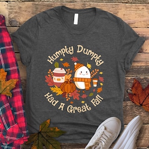 May include: Dark grey t-shirt with a fall-themed design. The graphic features the text "Humpty Dumpty Had A Great Fall" with an egg character wearing a scarf and hat, surrounded by leaves, a pumpkin, and coffee cups.