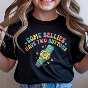 May include: A black t-shirt featuring a colorful graphic with the text SOME BELLIES HAVE TWO BUTTONS. The design includes a teal and yellow medical device illustration surrounded by small multi-colored hearts and stars in shades of pink, orange, and green.