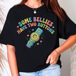 May include: Black t-shirt with the text "SOME BELLIES HAVE TWO BUTTONS" in rainbow colors. The graphic includes a cartoon remote control with a yellow circle and blue accents, surrounded by hearts.